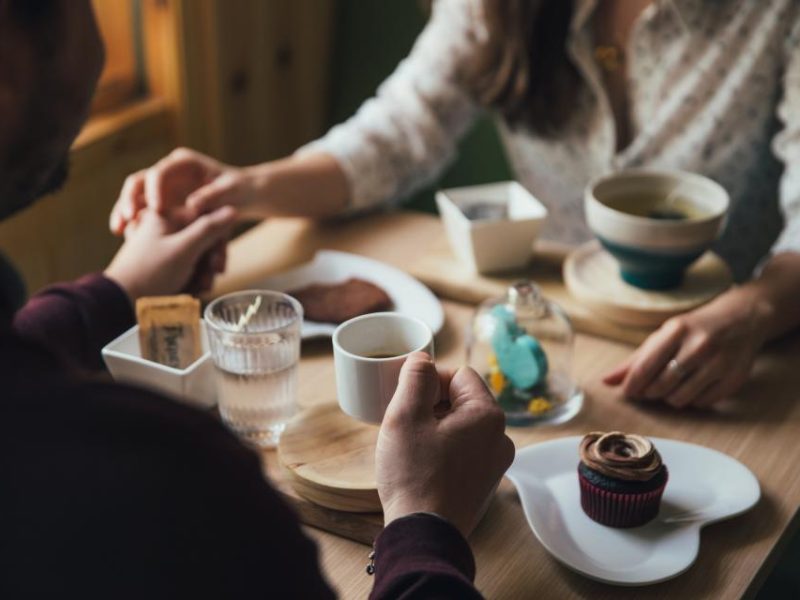 Two people holding hands across a cafe table with coffee and a cupcake