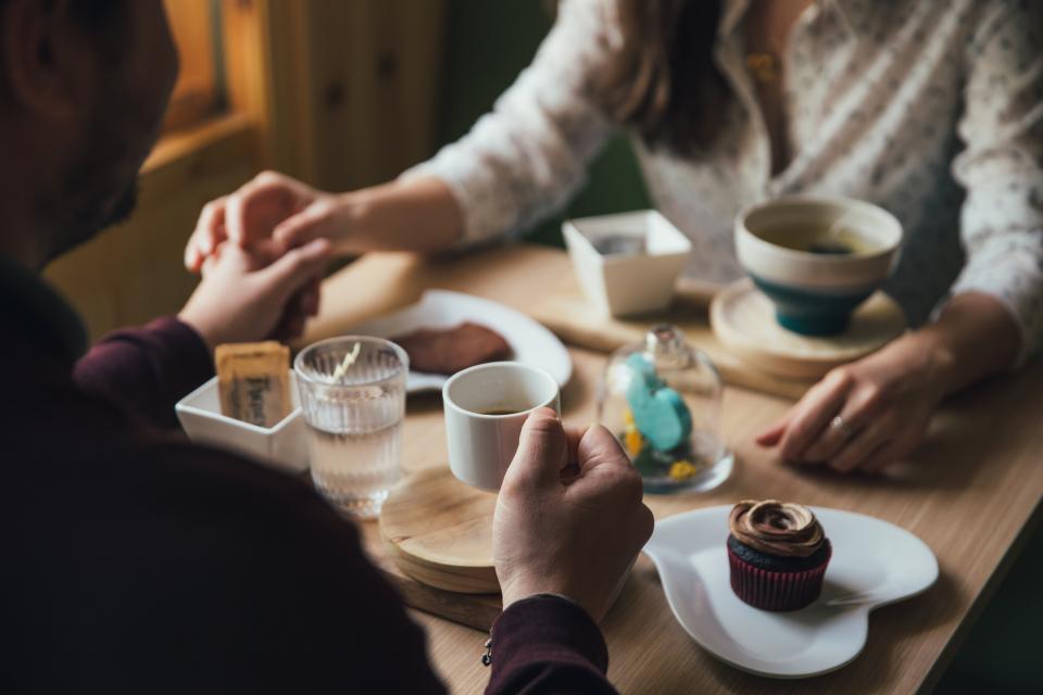 Two people holding hands across a cafe table with coffee and a cupcake